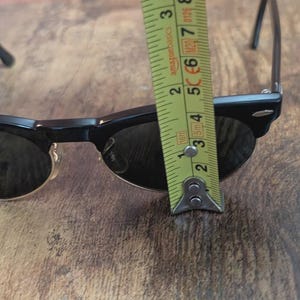 May include: Black sunglasses with a classic design, measured with a tape measure. The sunglasses are approximately 2 inches (5 cm) wide. The tape measure is yellow with black markings.