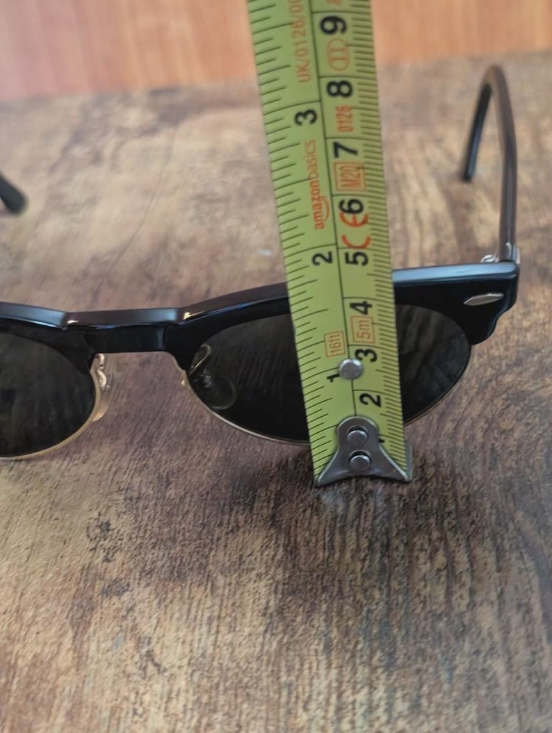 May include: Black sunglasses with a classic design, measured with a tape measure. The sunglasses are approximately 2 inches (5 cm) wide. The tape measure is yellow with black markings.