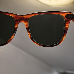 May include: Close-up of sunglasses with a tortoiseshell frame and dark lenses. The sunglasses have a classic, rectangular shape. The frame is a mix of brown and amber colors.