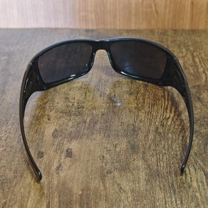 K&ouml;nnte beinhalten: Schwarze Sonnenbrille mit dunklen Gl&auml;sern. Die Sonnenbrille hat einen rechteckigen Rahmen und gebogene B&uuml;gel. Die Sonnenbrille liegt auf einer Holzoberfl&auml;che.
