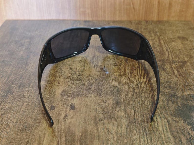 K&ouml;nnte beinhalten: Schwarze Sonnenbrille mit dunklen Gl&auml;sern. Die Sonnenbrille hat einen rechteckigen Rahmen und gebogene B&uuml;gel. Die Sonnenbrille liegt auf einer Holzoberfl&auml;che.