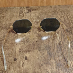 May include: Gold-framed sunglasses with dark lenses. The sunglasses have a unique octagonal shape and thin gold arms. The lenses appear to be a dark shade, providing a contrast to the gold frame. The arms have a clear, curved design.