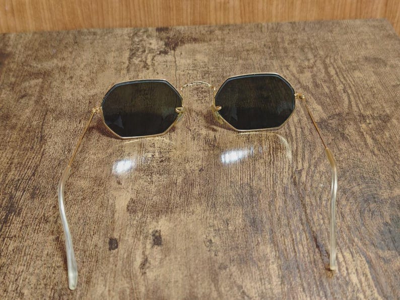 May include: Gold-framed sunglasses with dark lenses. The sunglasses have a unique octagonal shape and thin gold arms. The lenses appear to be a dark shade, providing a contrast to the gold frame. The arms have a clear, curved design.