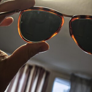 Puede incluir: Gafas de sol con montura de carey y lentes oscuras se sostienen a la luz. Las monturas tienen un dise&ntilde;o cl&aacute;sico con un c&aacute;lido patr&oacute;n marr&oacute;n y &aacute;mbar. Las lentes son de un color oscuro y s&oacute;lido, proporcionando sombra y protecci&oacute;n solar.