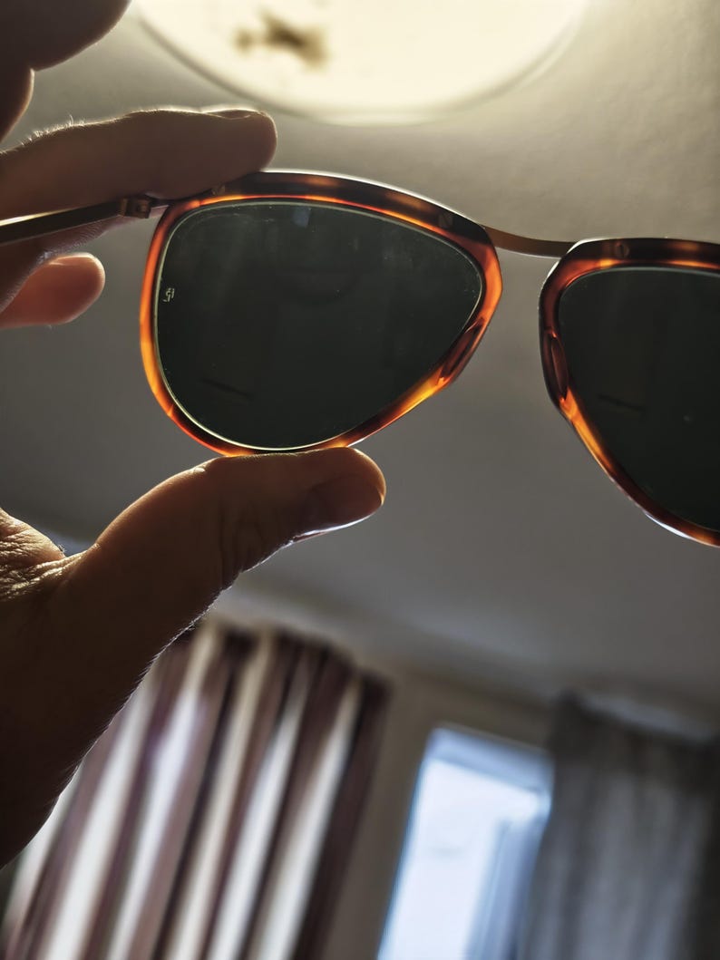 Puede incluir: Gafas de sol con montura de carey y lentes oscuras se sostienen a la luz. Las monturas tienen un dise&ntilde;o cl&aacute;sico con un c&aacute;lido patr&oacute;n marr&oacute;n y &aacute;mbar. Las lentes son de un color oscuro y s&oacute;lido, proporcionando sombra y protecci&oacute;n solar.