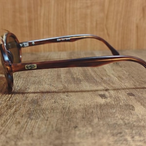 May include: Brown tortoiseshell sunglasses with dark lenses. The sunglasses have a rounded frame and a classic design. The arms of the sunglasses are a darker brown color. The sunglasses are resting on a wooden surface.