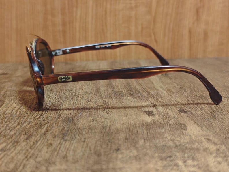 May include: Brown tortoiseshell sunglasses with dark lenses. The sunglasses have a rounded frame and a classic design. The arms of the sunglasses are a darker brown color. The sunglasses are resting on a wooden surface.