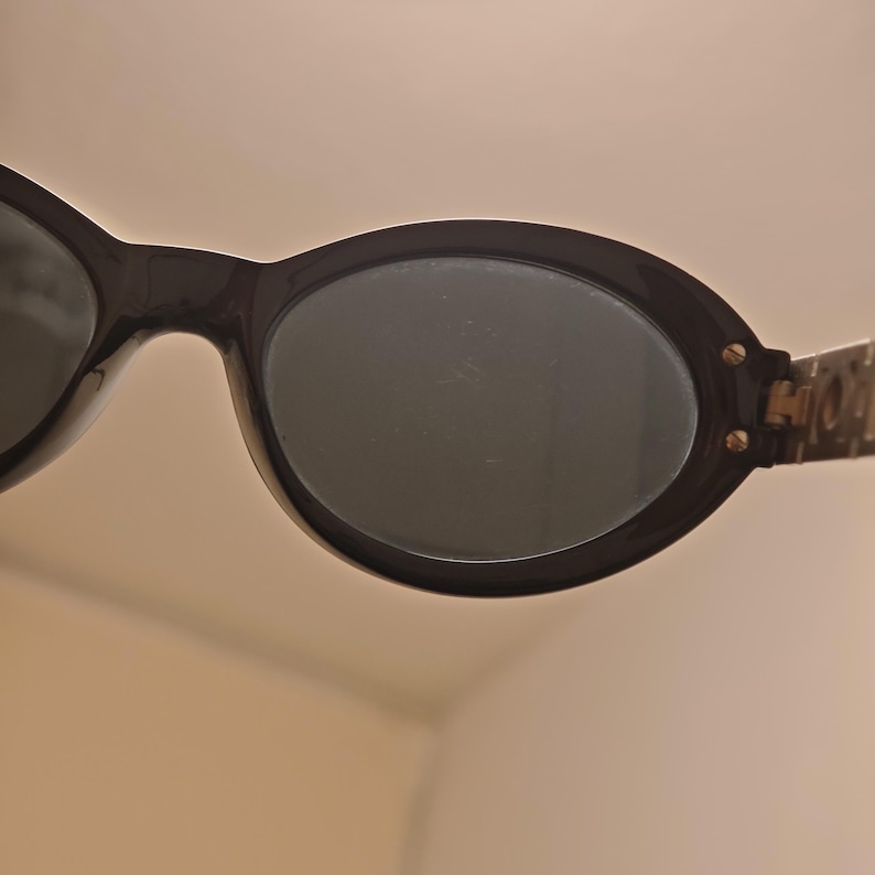 Puede incluir: Gafas de sol de forma ovalada con montura negra brillante y lentes oscuras. La montura presenta dos peque&ntilde;os tornillos met&aacute;licos en el borde exterior, donde la patilla se conecta con la parte frontal, aportando un detalle funcional.