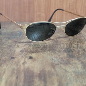 May include: Gold-toned sunglasses with black lenses. The frames have a sleek, retro design. The sunglasses are resting on a wooden surface. The Ray-Ban logo is visible on the lens.