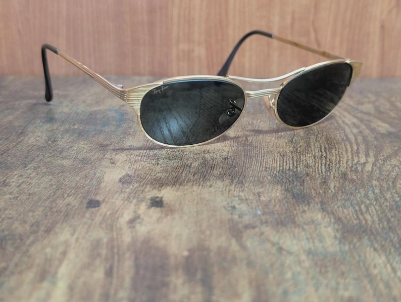 May include: Gold-toned sunglasses with black lenses. The frames have a sleek, retro design. The sunglasses are resting on a wooden surface. The Ray-Ban logo is visible on the lens.