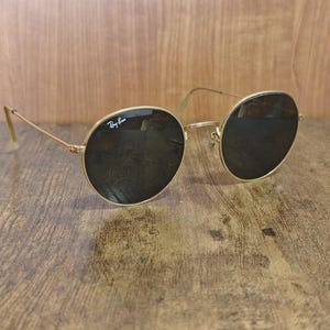 May include: Gold-framed sunglasses with dark lenses. The sunglasses have a classic round shape and the Ray-Ban logo is visible on the upper left lens. The glasses are resting on a wooden surface.