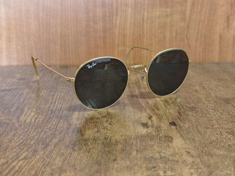 May include: Gold-framed sunglasses with dark lenses. The sunglasses have a classic round shape and the Ray-Ban logo is visible on the upper left lens. The glasses are resting on a wooden surface.