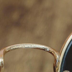 May include: Close-up of a gold-toned metal eyeglass frame. The text "RAY-BAN USA 54" is etched on the frame. The lens is black. The background is a blurred, warm brown.