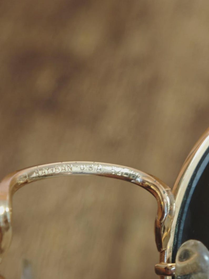 May include: Close-up of a gold-toned metal eyeglass frame. The text "RAY-BAN USA 54" is etched on the frame. The lens is black. The background is a blurred, warm brown.