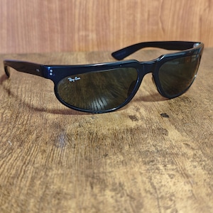 May include: Black sunglasses with dark lenses. The sunglasses have a rounded frame and a curved design. The Ray-Ban logo is visible on the left lens. The sunglasses are resting on a wooden surface.