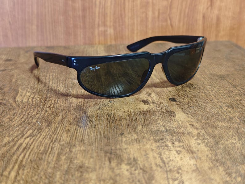May include: Black sunglasses with dark lenses. The sunglasses have a rounded frame and a curved design. The Ray-Ban logo is visible on the left lens. The sunglasses are resting on a wooden surface.