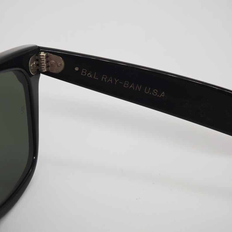 May include: Close-up of black sunglasses with green lenses. The text "B&L RAY-BAN U.S.A." is printed on the arm of the sunglasses. The hinge is visible, connecting the frame to the arm.