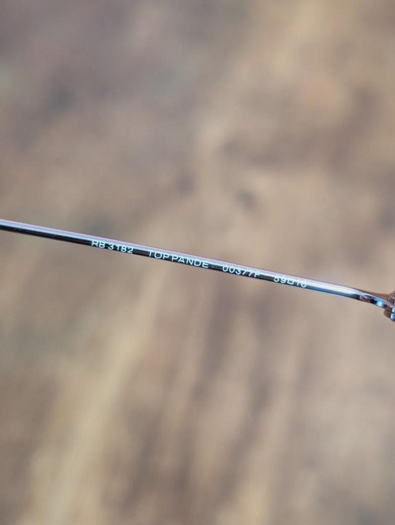 May include: Close-up of a silver metal eyeglass arm. The text "RB 3182 TOP PANDE 0037/F 59[]16" is printed in white on the arm. The background is a blurred, neutral tone.