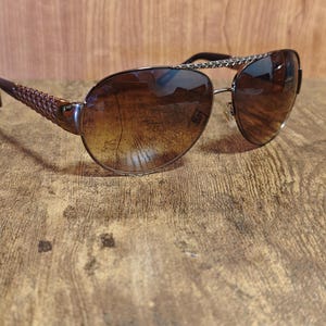 Könnte beinhalten: Braune Aviator-Sonnenbrille mit Farbverlaufsgläsern. Die Fassungen sind metallfarben mit einer dekorativen Kette oben. Die Bügel haben eine gewebte Textur. Die Sonnenbrille liegt auf einer Holzoberfläche.