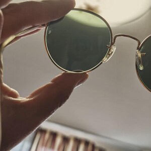 May include: Round sunglasses with gold-colored frames and green lenses are held up. The glasses have a classic design with a thin frame and a small logo on the lens. The lenses are a dark green color.
