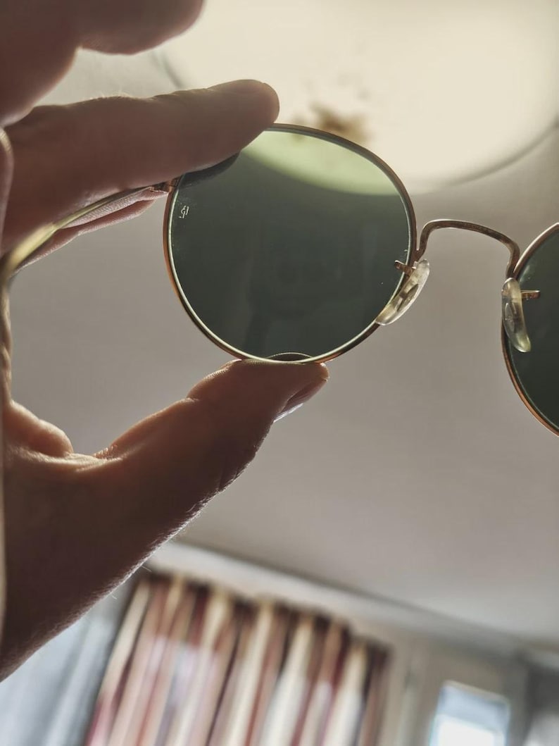 May include: Round sunglasses with gold-colored frames and green lenses are held up. The glasses have a classic design with a thin frame and a small logo on the lens. The lenses are a dark green color.