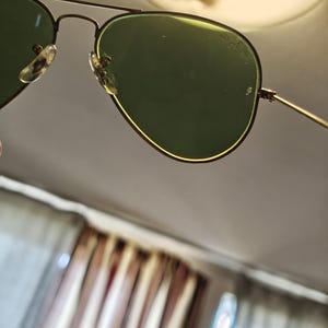 May include: Aviator sunglasses with green lenses and a gold-colored frame. The lenses have a slight curve, and the frame features a double bridge design. The nose pads are clear. The sunglasses are angled, with a blurred background.