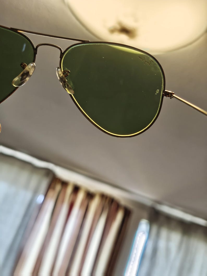 May include: Aviator sunglasses with green lenses and a gold-colored frame. The lenses have a slight curve, and the frame features a double bridge design. The nose pads are clear. The sunglasses are angled, with a blurred background.