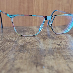 May include: Eyeglasses with a colorful striped pattern on the frames. The frames are primarily blue, with accents of black, red, and yellow. The lenses are clear, and the glasses are set against a wooden surface.