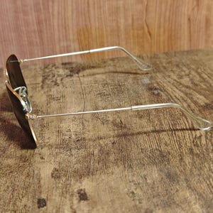 Puede incluir: Gafas de sol estilo aviador con montura de metal dorado y lentes oscuras. Las gafas de sol est&aacute;n sobre una superficie de madera, mostrando su dise&ntilde;o elegante y forma cl&aacute;sica.