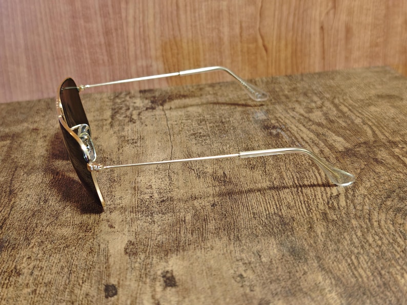 Puede incluir: Gafas de sol estilo aviador con montura de metal dorado y lentes oscuras. Las gafas de sol est&aacute;n sobre una superficie de madera, mostrando su dise&ntilde;o elegante y forma cl&aacute;sica.