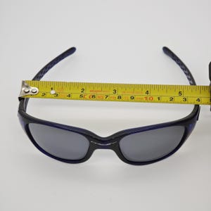 May include: Black and blue sunglasses with grey lenses. The sunglasses are measured with a yellow tape measure, showing a width of approximately 14 cm. The brand name "Oakley" is visible on the frame.