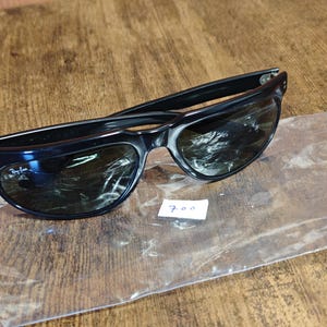 May include: Black sunglasses with dark lenses, resting on a wooden surface. The sunglasses are in a clear plastic bag. A small white tag with the number "700" is visible.