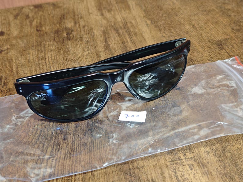 May include: Black sunglasses with dark lenses, resting on a wooden surface. The sunglasses are in a clear plastic bag. A small white tag with the number "700" is visible.
