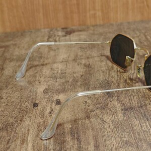 May include: Gold-rimmed sunglasses with dark lenses. The frames have a unique octagonal shape. The temples are silver and have clear plastic earpieces. The sunglasses are resting on a wooden surface.