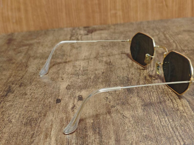 May include: Gold-rimmed sunglasses with dark lenses. The frames have a unique octagonal shape. The temples are silver and have clear plastic earpieces. The sunglasses are resting on a wooden surface.