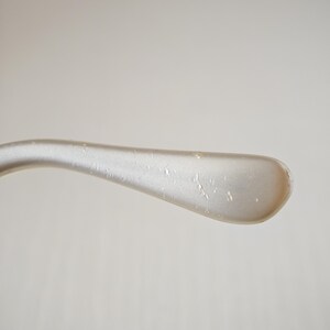 May include: A curved, light-colored temple arm of a pair of eyeglasses. The surface has a smooth, pearlescent finish with a subtle sheen and several small, scattered indentations or marks visible along the length of the curved plastic material.