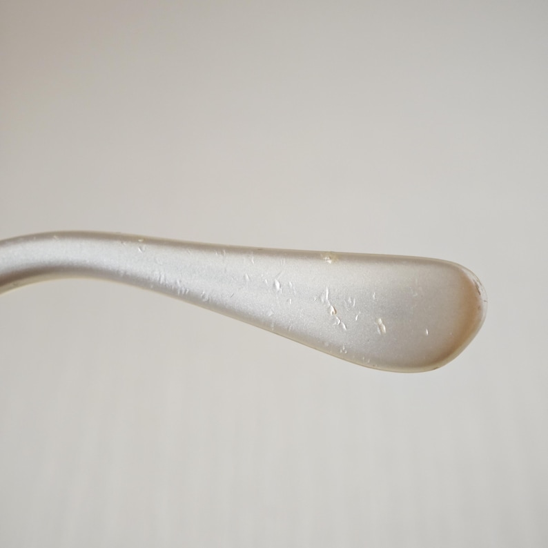 May include: A curved, light-colored temple arm of a pair of eyeglasses. The surface has a smooth, pearlescent finish with a subtle sheen and several small, scattered indentations or marks visible along the length of the curved plastic material.
