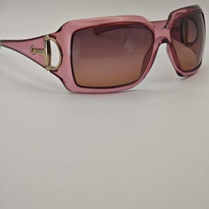 May include: Pink-framed sunglasses with gradient brown lenses. The sunglasses have a square shape and a gold-colored accent on the temples with the brand name. The lenses transition from a darker shade at the top to a lighter shade at the bottom.