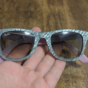Puede incluir: Gafas de sol Wayfarer grises con un patr&oacute;n veteado y patillas rosas. El logotipo de Ray-Ban es visible en la lente izquierda. Las gafas tienen lentes oscuras.