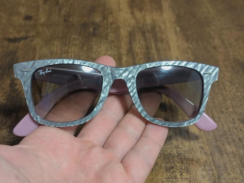 Puede incluir: Gafas de sol Wayfarer grises con un patr&oacute;n veteado y patillas rosas. El logotipo de Ray-Ban es visible en la lente izquierda. Las gafas tienen lentes oscuras.