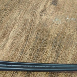 May include: A black, elongated object with text etched into its surface, resting on a textured, brown wooden surface. The object appears to be a tool or part of a larger mechanism.