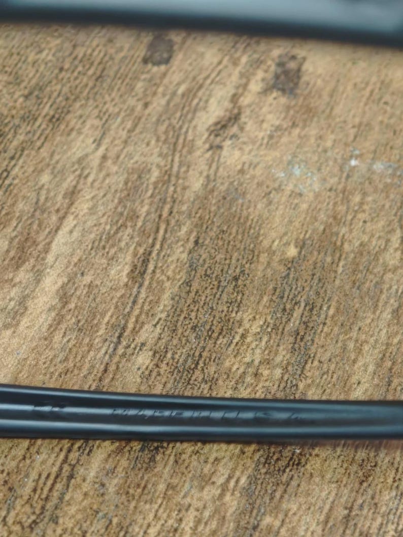 May include: A black, elongated object with text etched into its surface, resting on a textured, brown wooden surface. The object appears to be a tool or part of a larger mechanism.