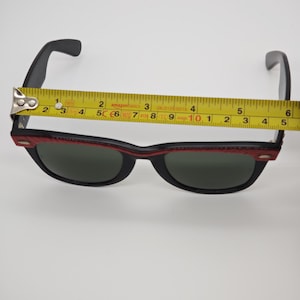 May include: Black sunglasses with a red and black frame. The sunglasses are measured with a tape measure, showing a length of approximately 14 cm. The lenses are dark green.