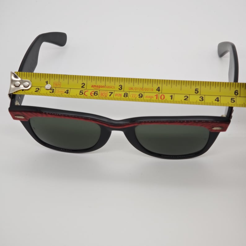 May include: Black sunglasses with a red and black frame. The sunglasses are measured with a tape measure, showing a length of approximately 14 cm. The lenses are dark green.
