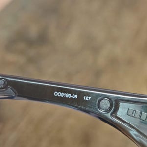 May include: Close-up of a section of eyeglasses with the text "OO9190-05 127" printed on the arm. The frame is a translucent gray color.