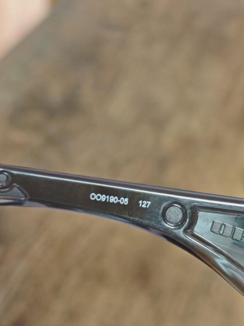 May include: Close-up of a section of eyeglasses with the text "OO9190-05 127" printed on the arm. The frame is a translucent gray color.