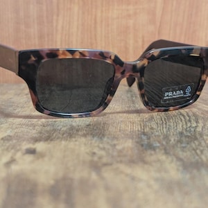 May include: Prada sunglasses with a tortoiseshell pattern frame and dark lenses. The sunglasses have a square shape and a sticker on the right lens. The arms are a solid brown color.
