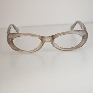 May include: A pair of translucent taupe-colored eyeglasses with an oval-shaped frame. The glasses feature a smooth, glossy finish and small metallic decorative accents on the outer corners of the frame near the hinges of the temples.