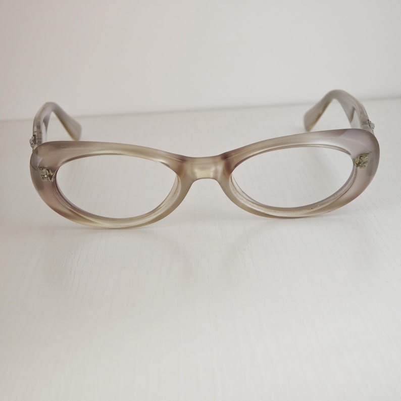 May include: A pair of translucent taupe-colored eyeglasses with an oval-shaped frame. The glasses feature a smooth, glossy finish and small metallic decorative accents on the outer corners of the frame near the hinges of the temples.