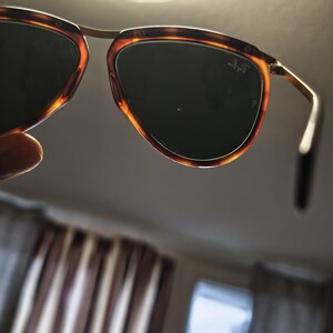 Puede incluir: Gafas de sol con montura de carey y lentes oscuras. Las gafas tienen un puente y varillas de metal dorado. El logotipo de Ray-Ban es visible en la lente.